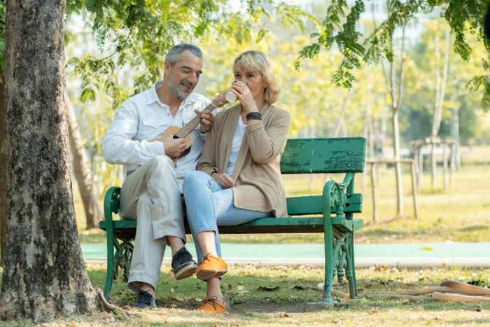 Happy senior man and woman on park bench, man plays ukulele