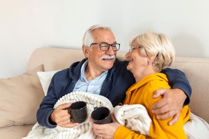 Loving elderly couple relaxing on the sofa with mugs and a cozy blanket, smiling warmly at each other