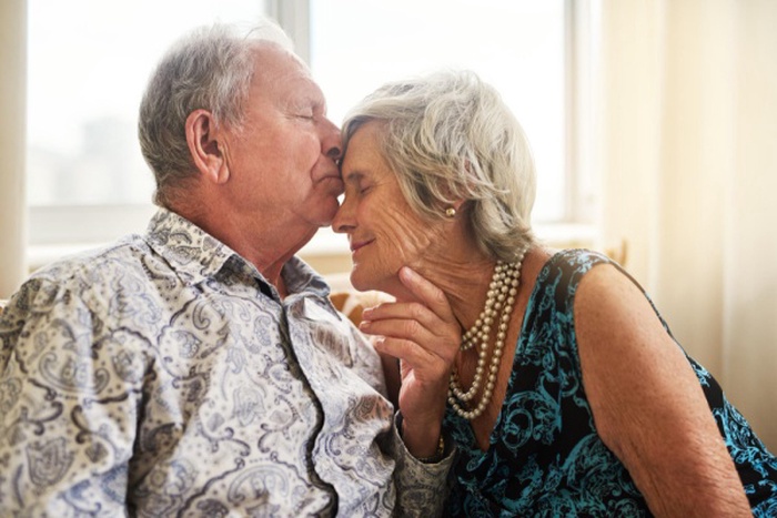 Senior couple and kiss at nursing home with love forehead and bonding in living room retirement care and relax in a house happy with elderly people on a sofa with marriage and support together