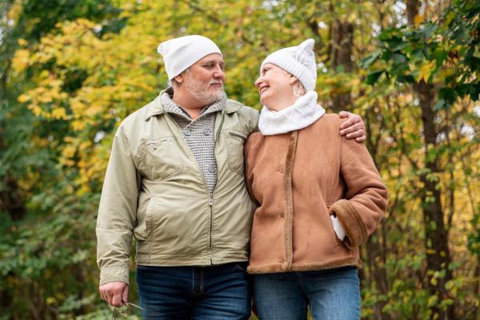 Loving older couple wearing winter beanies walking in autumn park, romantic elderly pair enjoying outdoor fall date