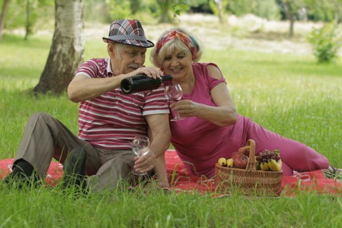 Happy elderly white couple enjoying outdoor picnic pouring red wine on green lawn, mature dating lifestyle for over 50 singles