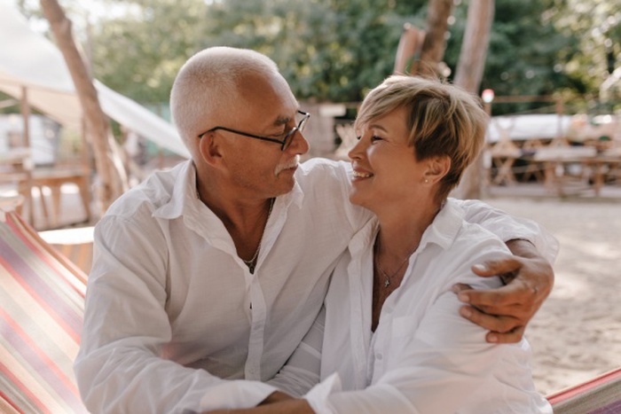Gray-haired man with eyeglasses in long sleeve shirt hugging and looking at short haired smiling wife in white blouse on beach.