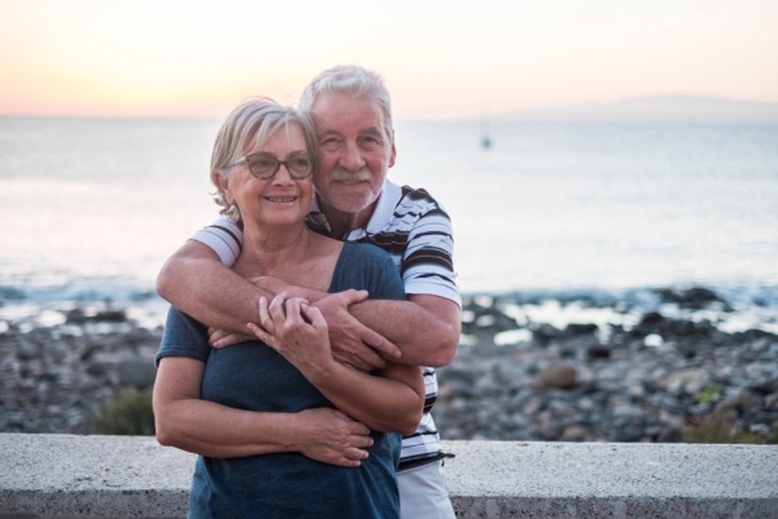 Couple of seniors hogging at the beach with a lot of love - retired together - woman with glasses and man with sea background